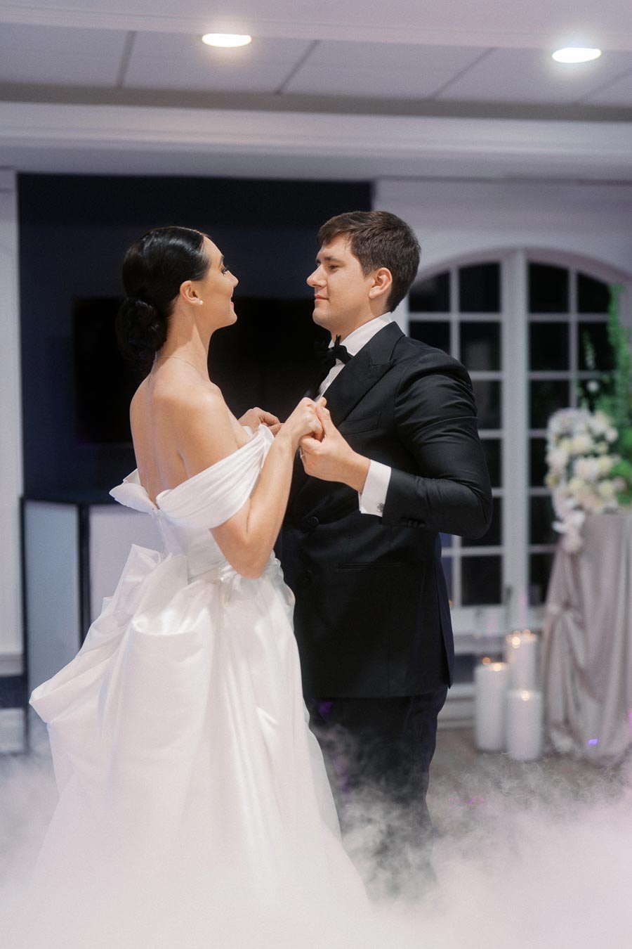 A bride and groom share a romantic first dance on their wedding day, surrounded by mist. The bride is wearing an elegant white gown, and the groom is in a black tuxedo, with candles and floral decorations enhancing the ambiance.