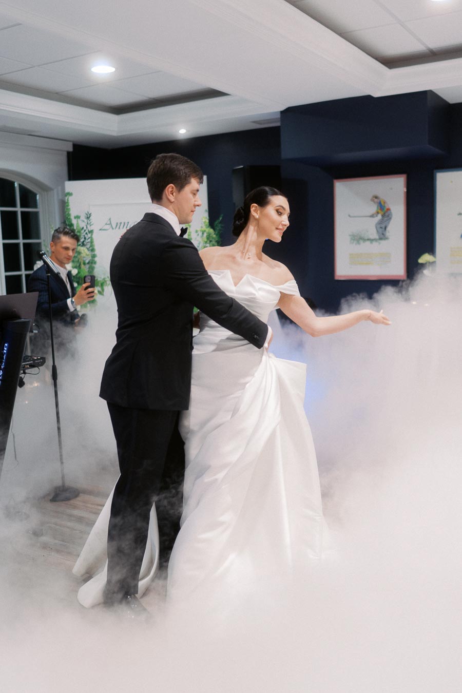 A bride and groom in elegant attire share a romantic first dance on a fog-covered floor, creating a dreamy wedding atmosphere indoors, while a guest captures the moment on their phone in the background.
