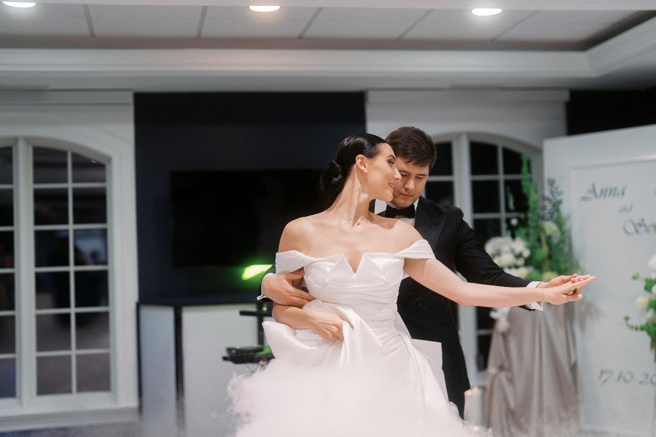 A bride and groom gracefully dancing at their wedding reception, with the bride wearing an elegant off-shoulder white dress and the groom in a classic black tuxedo, in a beautifully decorated venue.