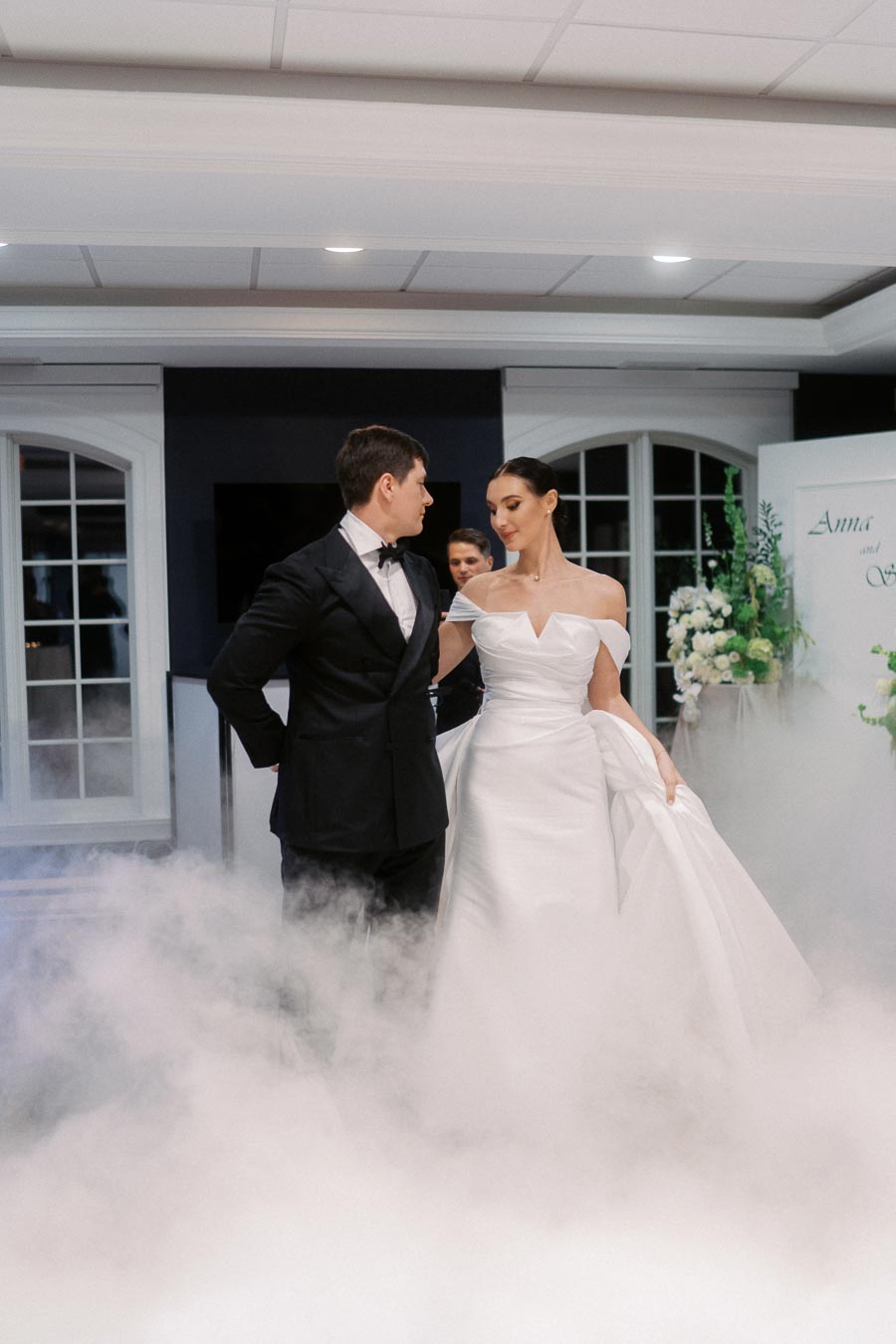 A newlywed couple elegantly dancing on a fog-covered floor, with the bride wearing a white off-shoulder wedding gown and the groom in a classic black tuxedo, creating a romantic atmosphere in a decorated venue.