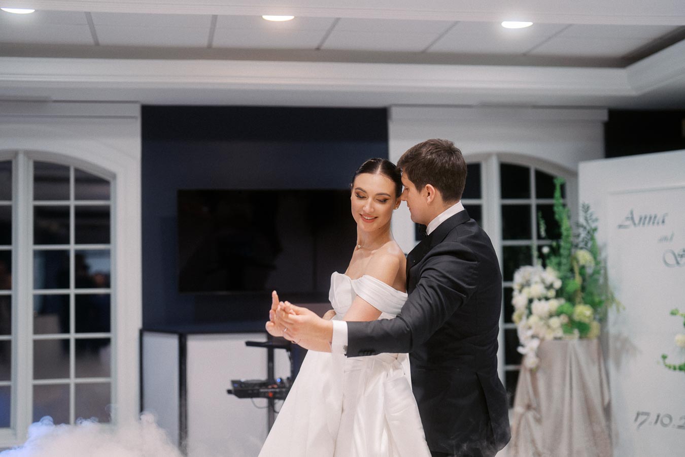 Bride and groom share a romantic first dance at wedding reception, elegantly dressed with floral decorations in background.
