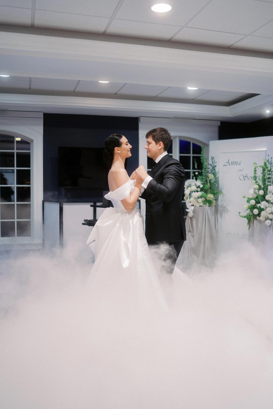 Elegant couple dancing at their wedding reception, surrounded by mist, in a beautifully decorated venue with floral arrangements.