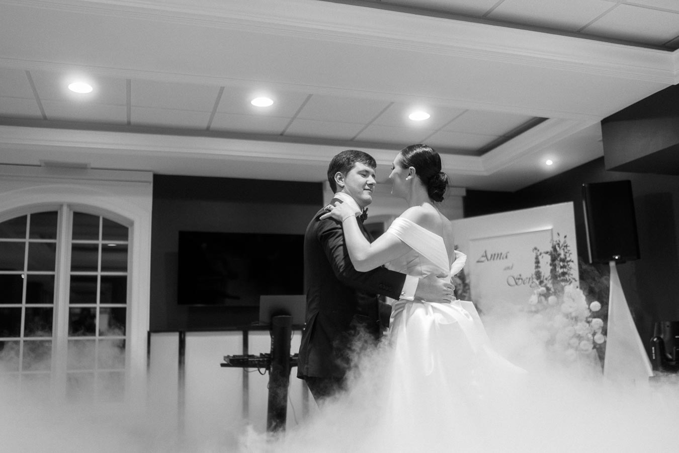 Wedding couple dancing on a smoke-covered floor in an elegantly decorated venue, capturing a romantic moment in black and white.