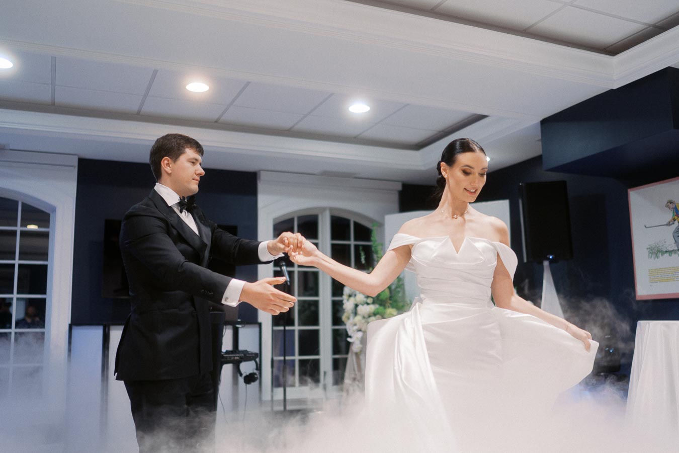 Elegant bride and groom performing their first dance in a beautifully lit venue with fog effects, showcasing the bride's stunning white gown and the groom's classic black tuxedo.