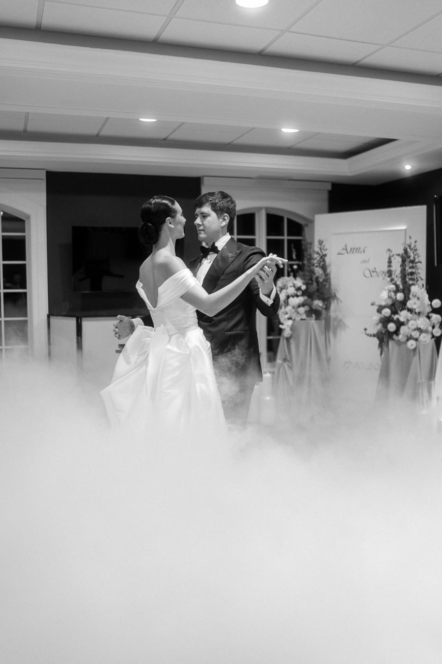 Black and white image of a couple dancing elegantly in a misty wedding venue, with the bride in an off-the-shoulder gown and the groom in a tuxedo, surrounded by floral arrangements.
