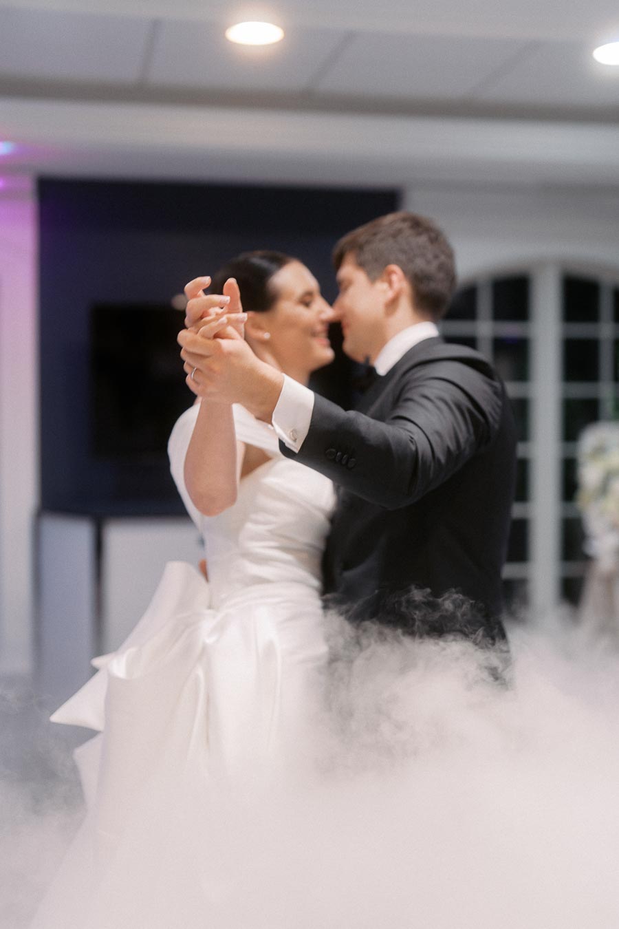 A newlywed couple shares their first dance surrounded by romantic smoke effects, the bride in an elegant white gown and the groom in a classic black suit, against a beautifully lit wedding venue backdrop.