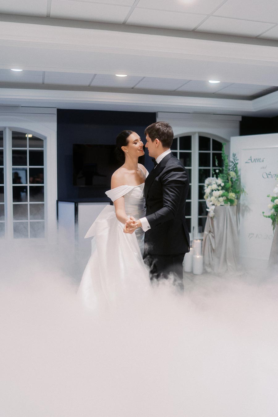Bride and groom dance on their wedding day surrounded by romantic mist, in a beautifully decorated venue with soft lighting and floral arrangements.