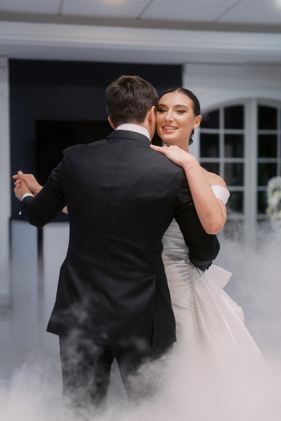 A bride and groom share their first dance amidst a cloud of mist, capturing a romantic moment during their wedding reception in a softly lit venue.