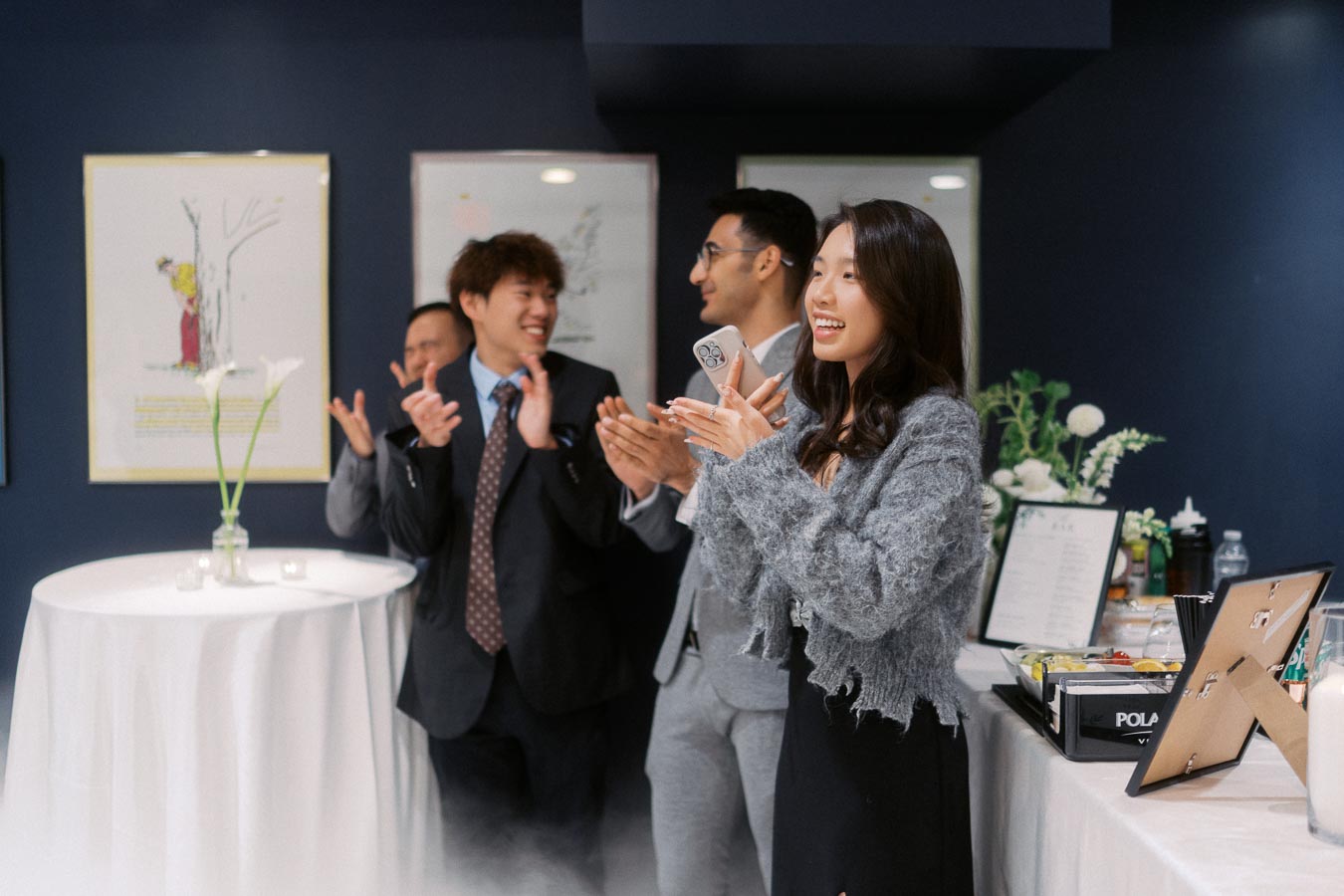 A group of people at a social event, with one woman in focus holding a smartphone and smiling, while others in the background are clapping and laughing, in a casually decorated room with flowers and artwork.
