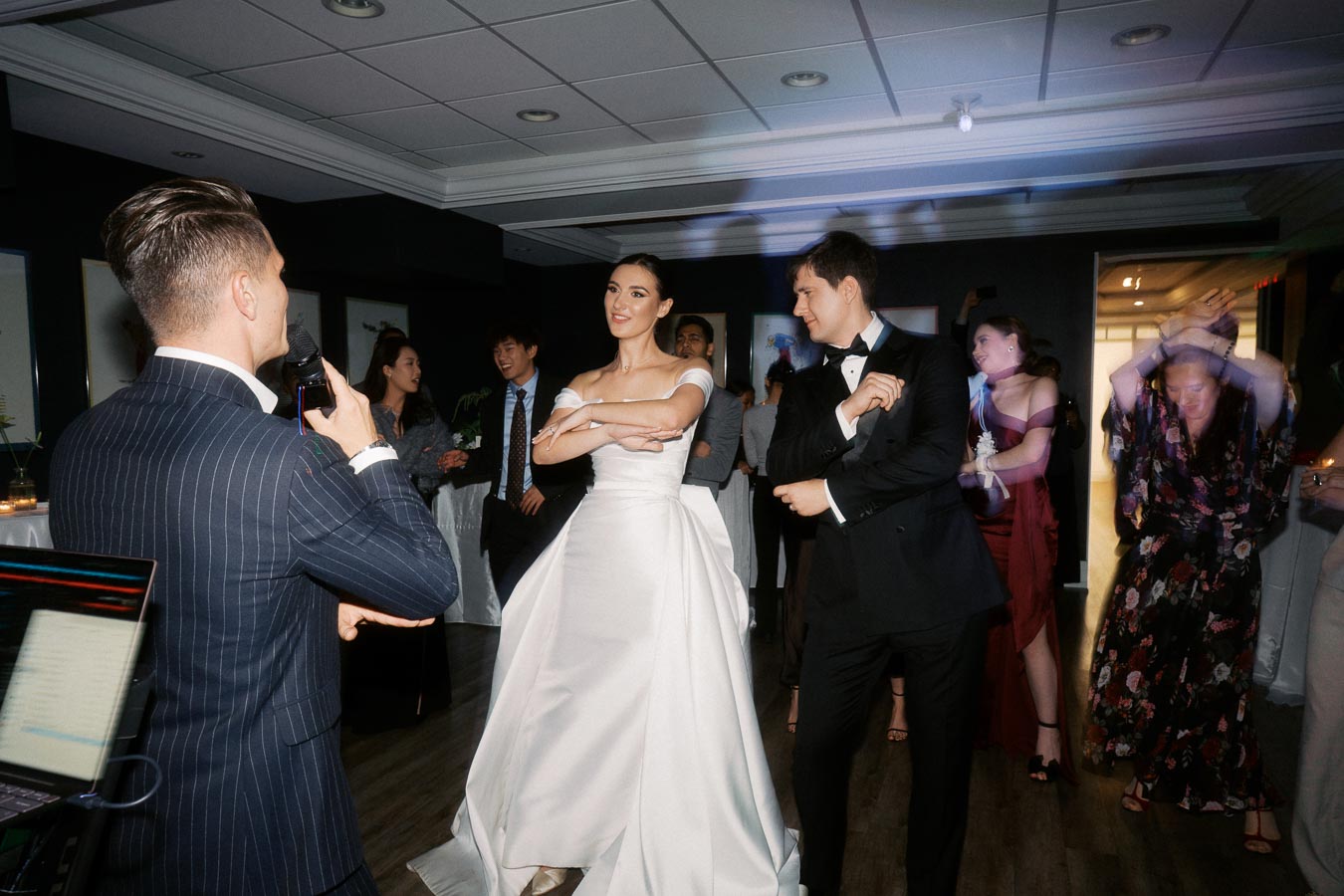 A joyful wedding reception scene with a bride in a white gown and groom in a tuxedo dancing while guests cheer, under soft lighting in an indoor venue.