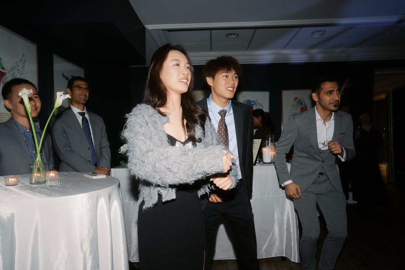 A group of well-dressed individuals engaging in a lively dance at a formal event, with tables adorned with flowers and candles in the background, creating a festive ambiance.