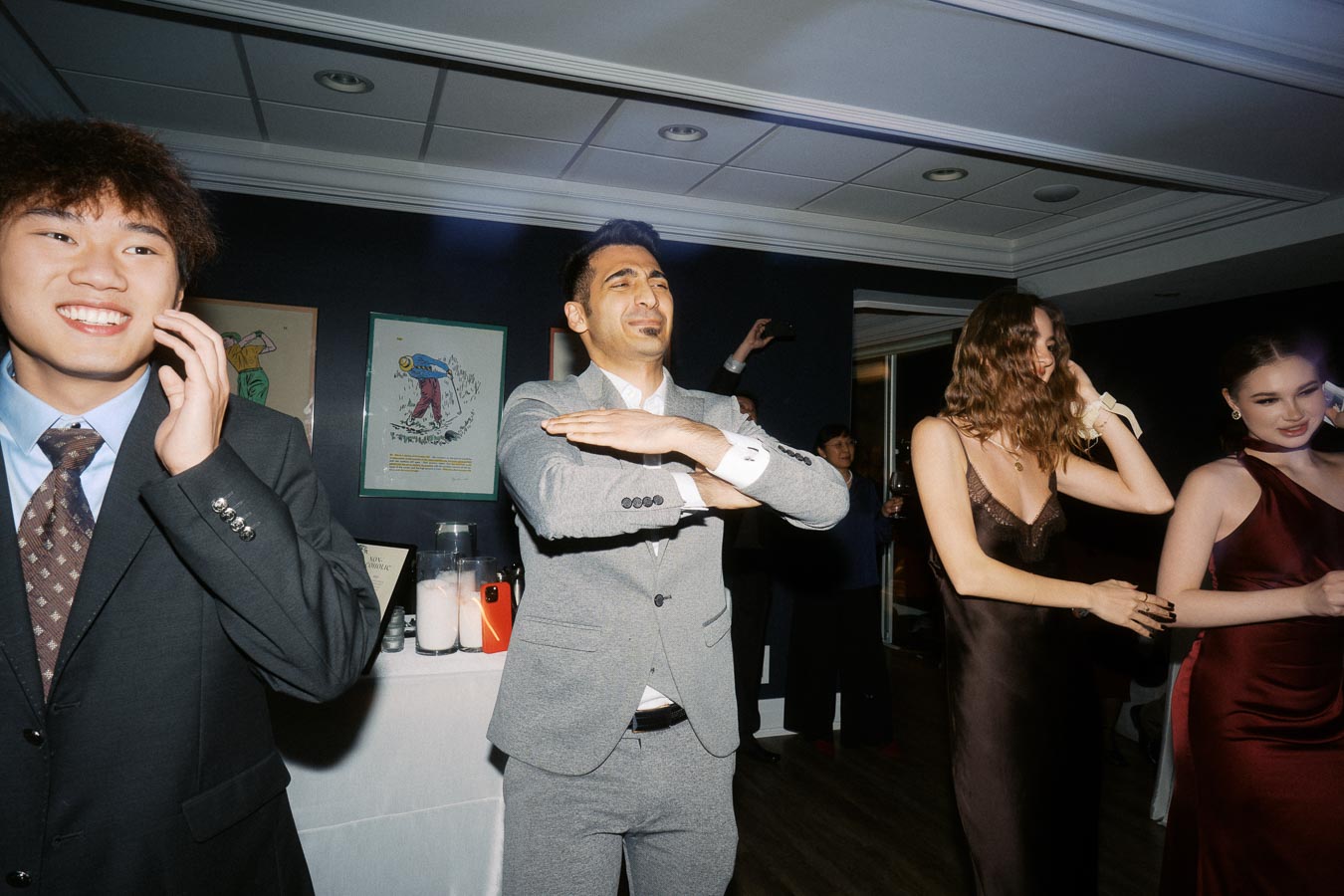 A group of people in formal attire enjoying a lively party indoors, with individuals laughing, talking on phones, and dancing in a dimly lit room decorated with artwork on the walls.
