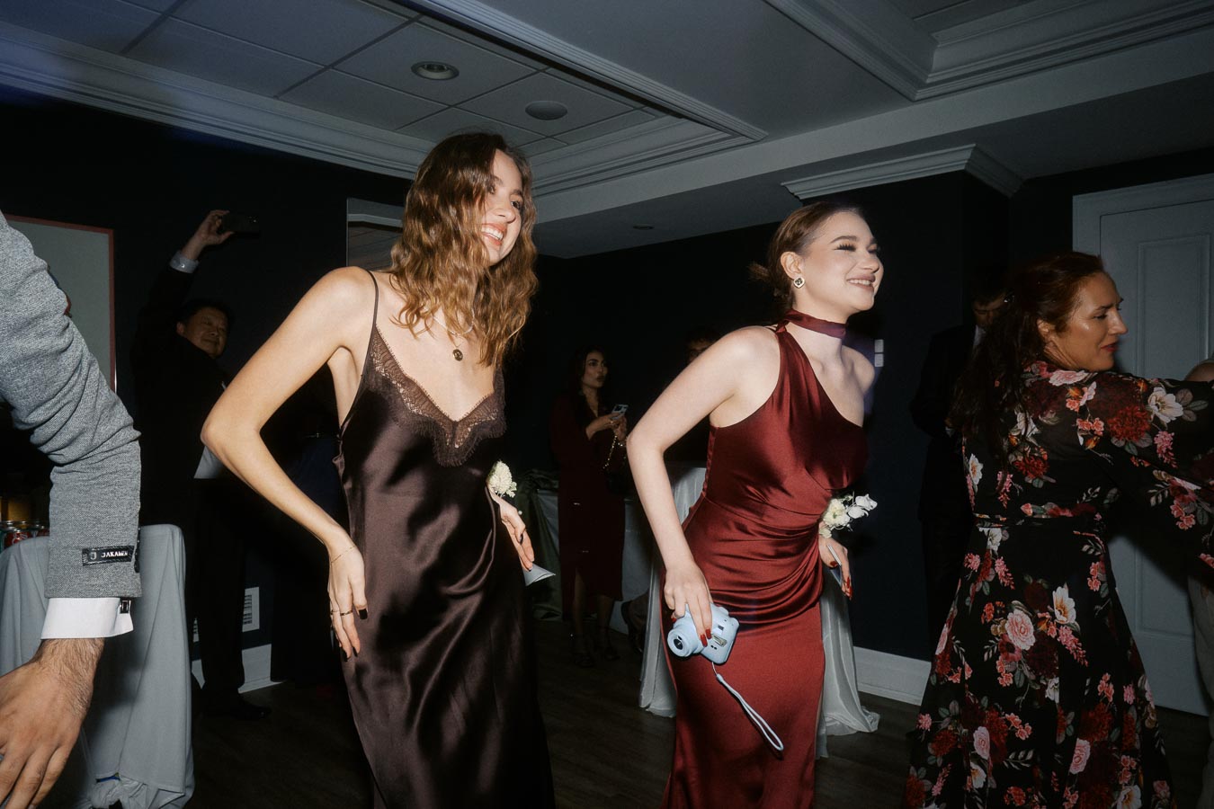 Group of people dancing joyfully at an indoor event, with a focus on two women in elegant dresses enjoying the moment.