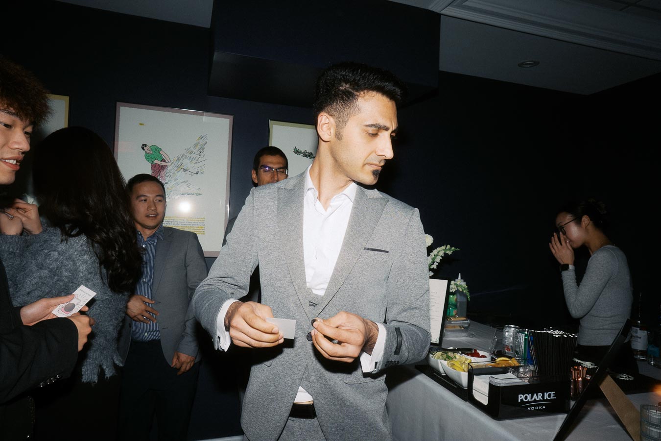 A group of people socializing at a formal event, with a man in a gray suit holding cards. A well-stocked bar and framed art are visible in the background, suggesting a sophisticated gathering.