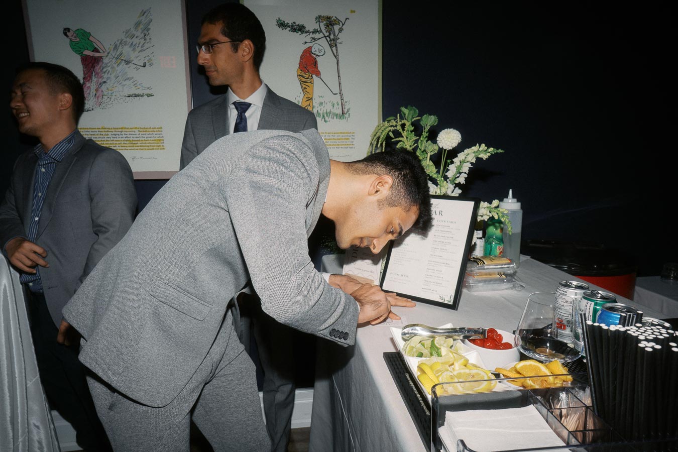 Three men in suits standing and interacting near a bar setup with a menu and various garnishes, in a dimly lit room with golf-themed artwork in the background.