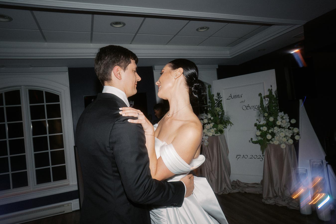 Bride and groom dancing closely at their wedding reception, surrounded by floral decorations and dim lighting, with a sign displaying Anna and Sergei and the date 17.10.2023 in the background.