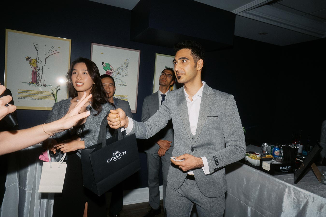 A group of well-dressed individuals at a social event, one handing out a Coach gift bag.