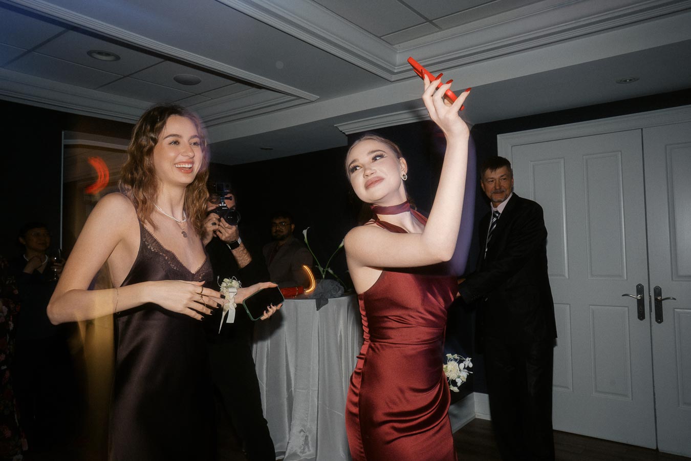 Two young women in elegant dresses enjoying a celebration, with one holding a smartphone and the other smiling, while a man in a suit observes in the background.