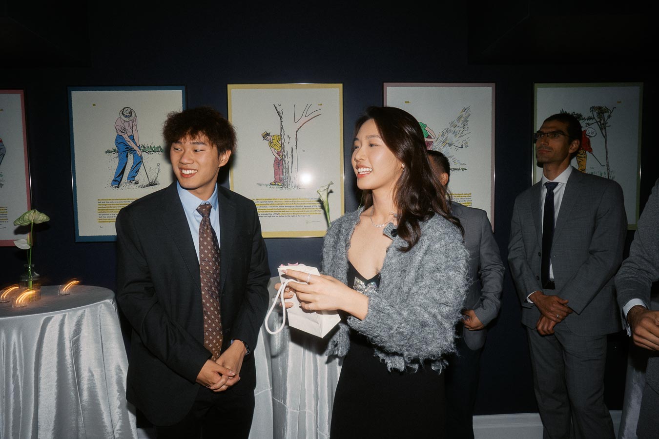 A group of elegantly dressed people attending an art exhibition with framed artwork on the dark walls, engaging in conversation and smiling.