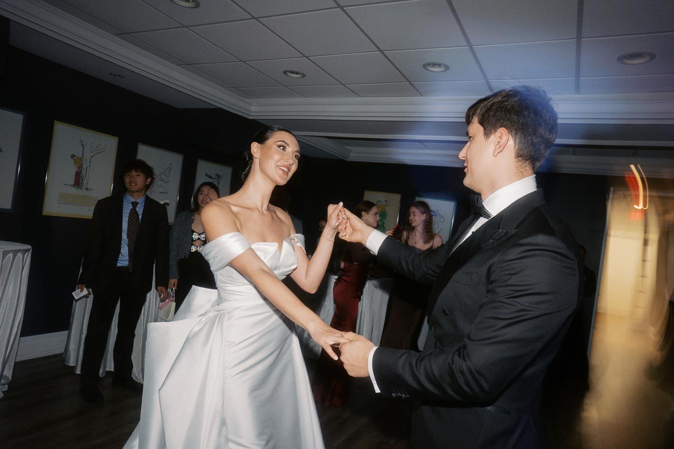 A smiling couple elegantly dancing at a formal event, with the woman in a stunning white wedding dress and the man in a classic black tuxedo, surrounded by a lively crowd in the background enjoying the celebration.