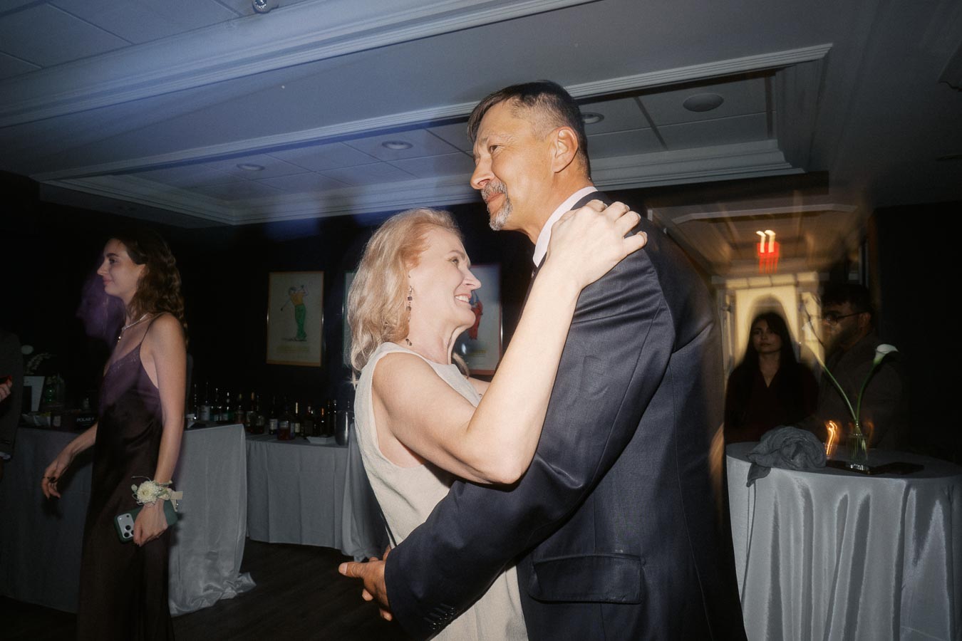 A couple dancing closely at an indoor event, with guests socializing in the background. The woman smiles as she embraces her partner, creating a warm and joyful atmosphere.