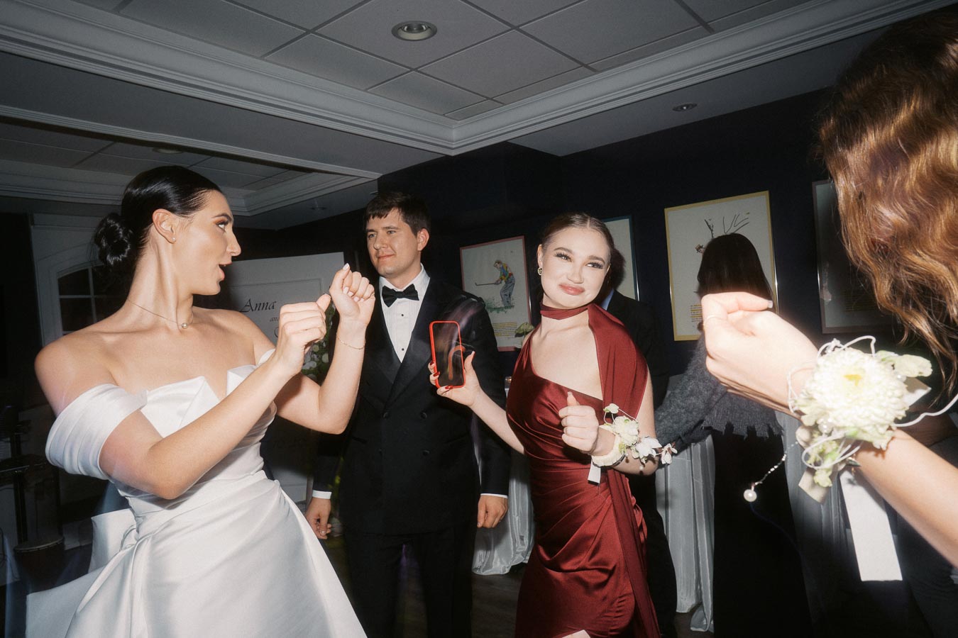 Group of elegantly dressed young adults dancing at a formal event, featuring a woman in a white gown and another in a red dress, displaying joyful expressions.