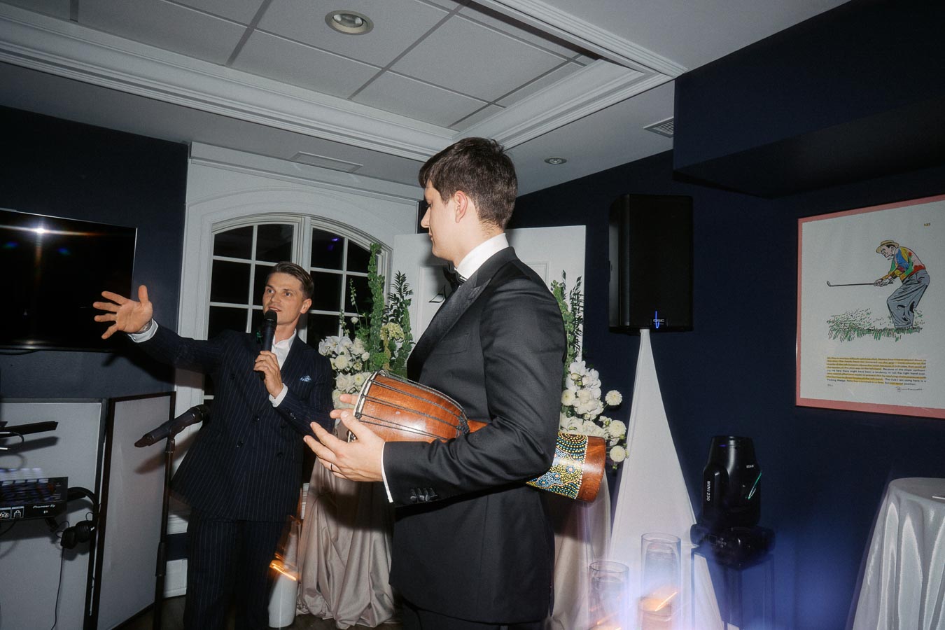 A speaker in a dark suit gestures with a microphone while engaging an audience at a formal event. Another person in a tuxedo stands nearby holding a decorated djembe drum. The room features stylish decor with white flowers and ambient lighting.
