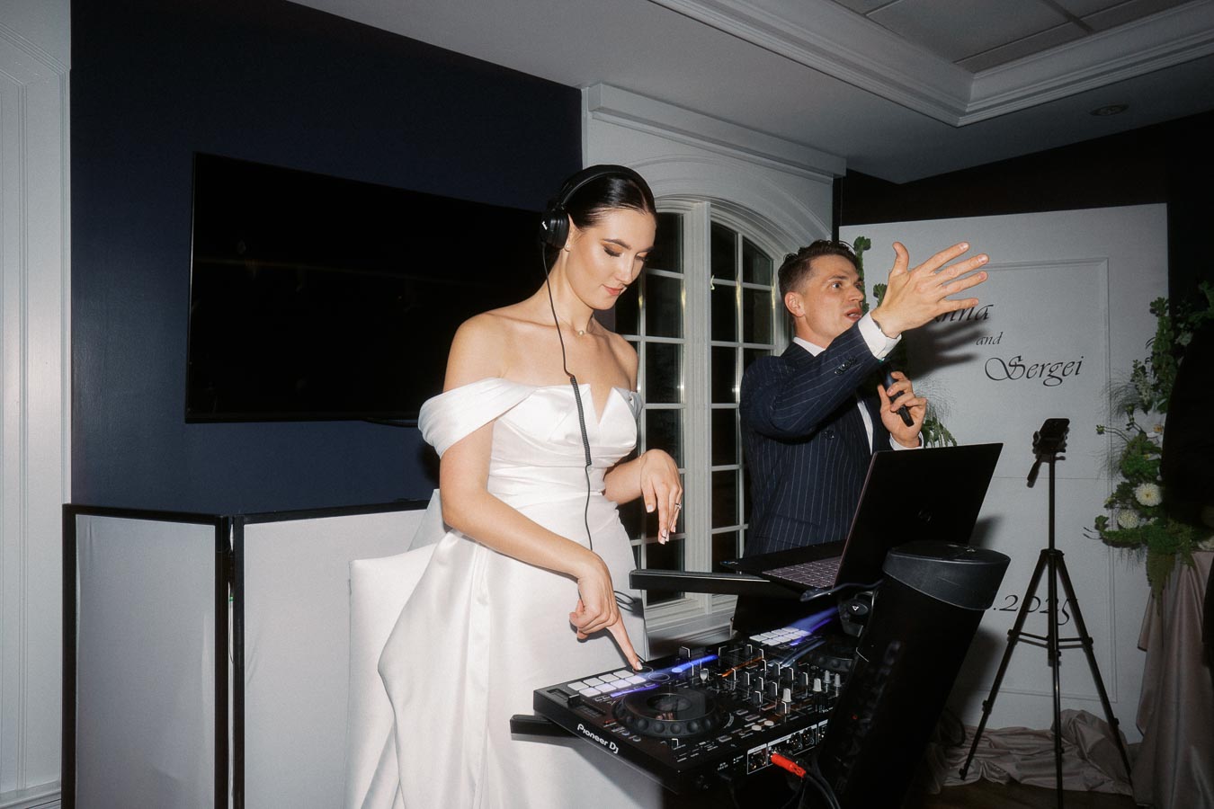 A woman in an elegant white dress DJing at a wedding reception with a man in a suit speaking into a microphone, with a sign that reads Anna and Sergei in the background.