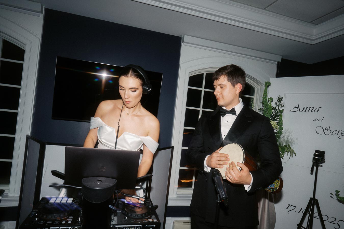 DJ playing music at an elegant event, accompanied by a man in a tuxedo holding a drum, set against a backdrop with a floral arrangement and personalized decor.