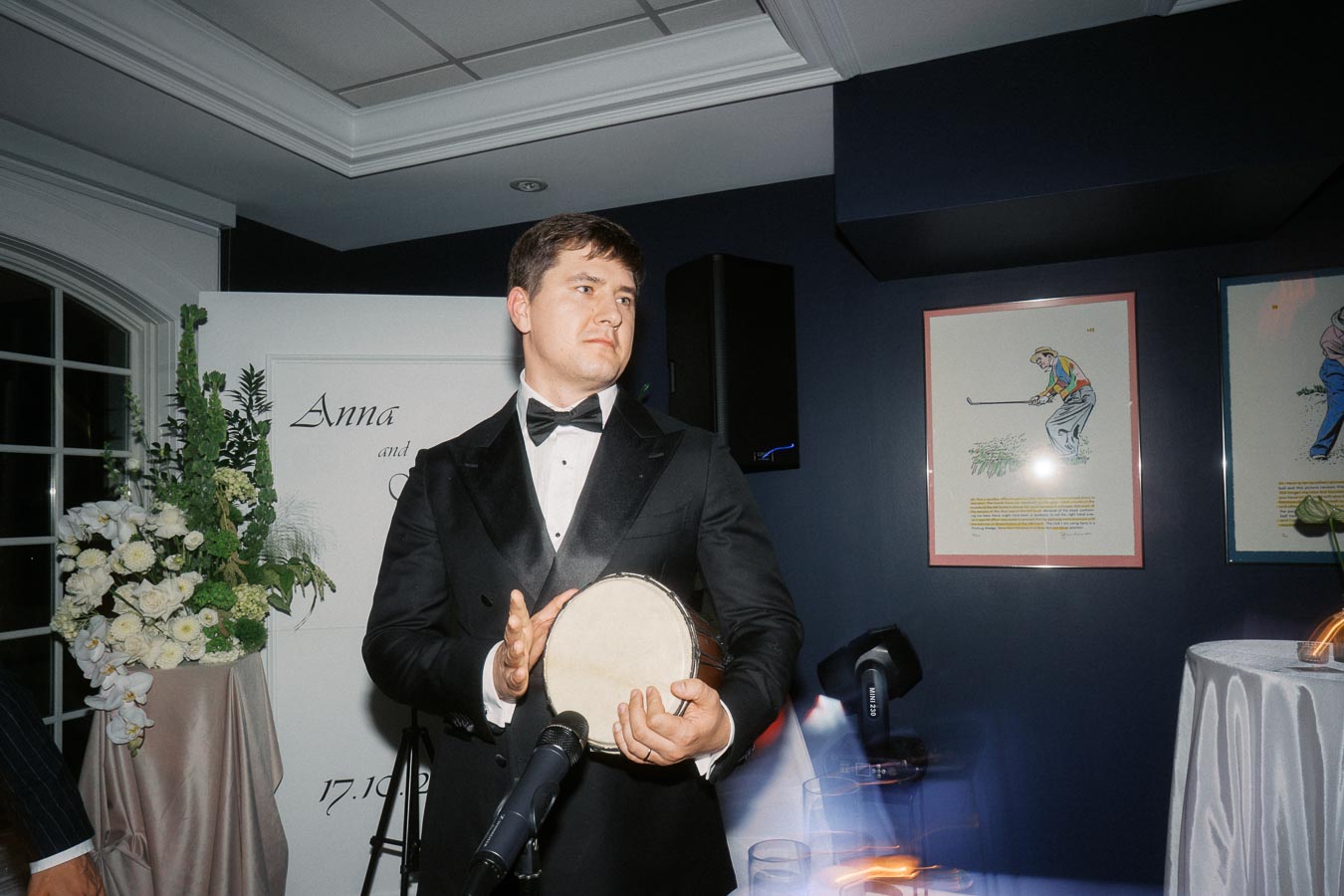 A man in a tuxedo plays a hand drum at a formal event, standing near a microphone with floral arrangements and artwork in the background.