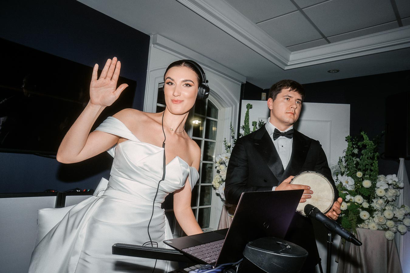 Bride DJ wearing headphones and a white dress waves while standing beside a groom in a tuxedo holding a small drum, with a laptop and microphone in front of them, set in an indoor venue with floral decorations.