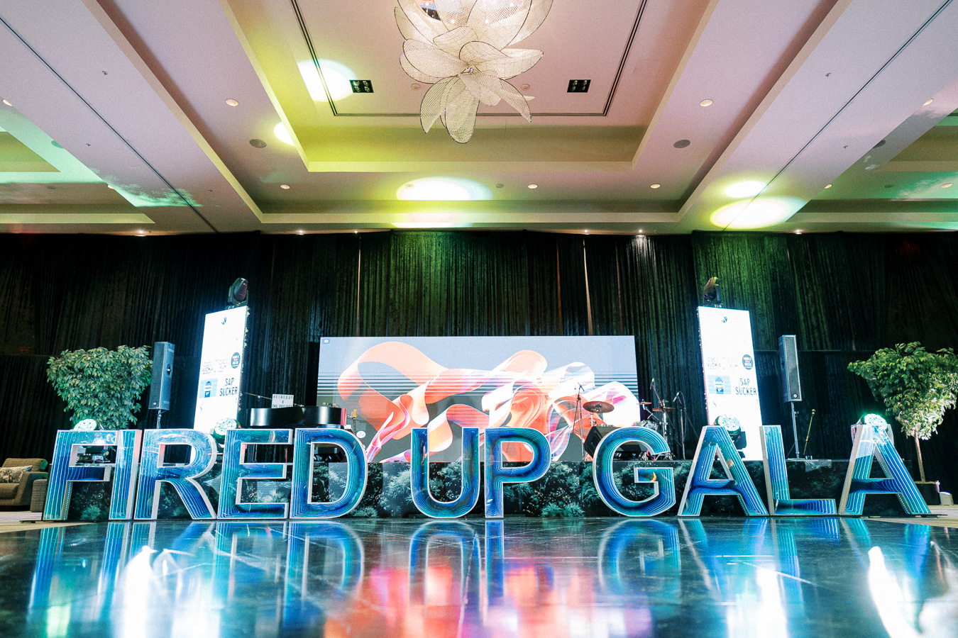 Elegant event stage set up for the Fired Up Gala, featuring large illuminated letters on a shiny floor, greenery, and a modern abstract backdrop under sophisticated ceiling lighting.