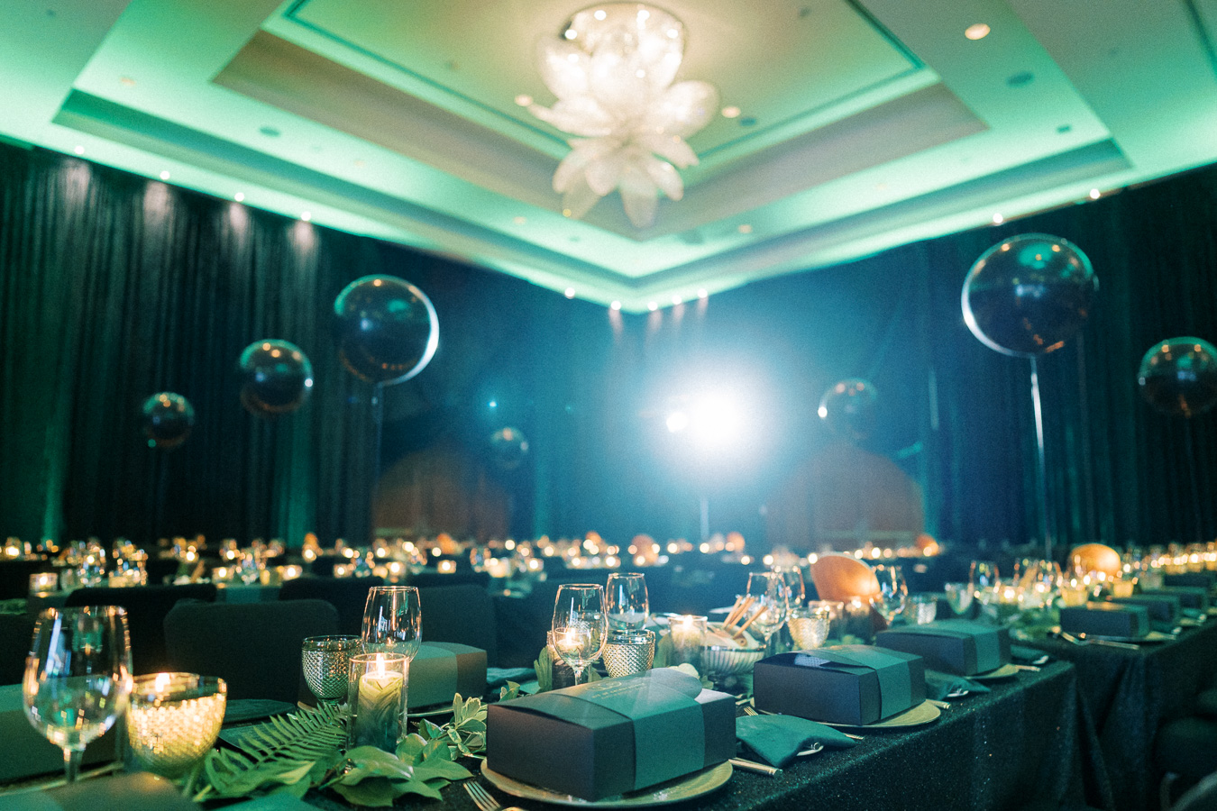 Elegant banquet hall setup with glowing candlelit tables, decorative black balloons, and luxurious place settings under a large chandelier, creating an ambient atmosphere for a formal event.