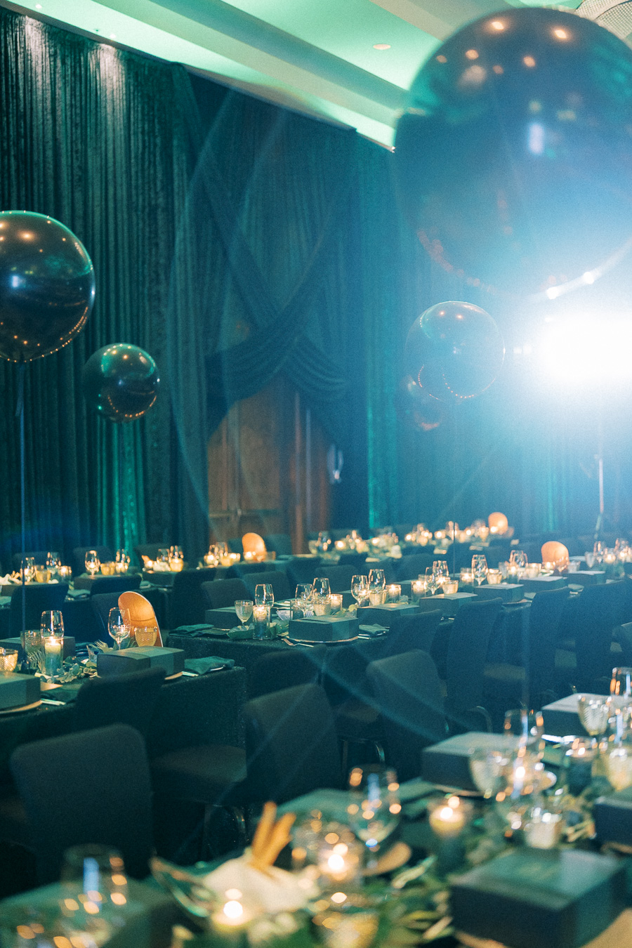 Elegant dining setup for an event with long banquet tables decorated with candles and glassware, surrounded by large metallic balloons under soft lighting.