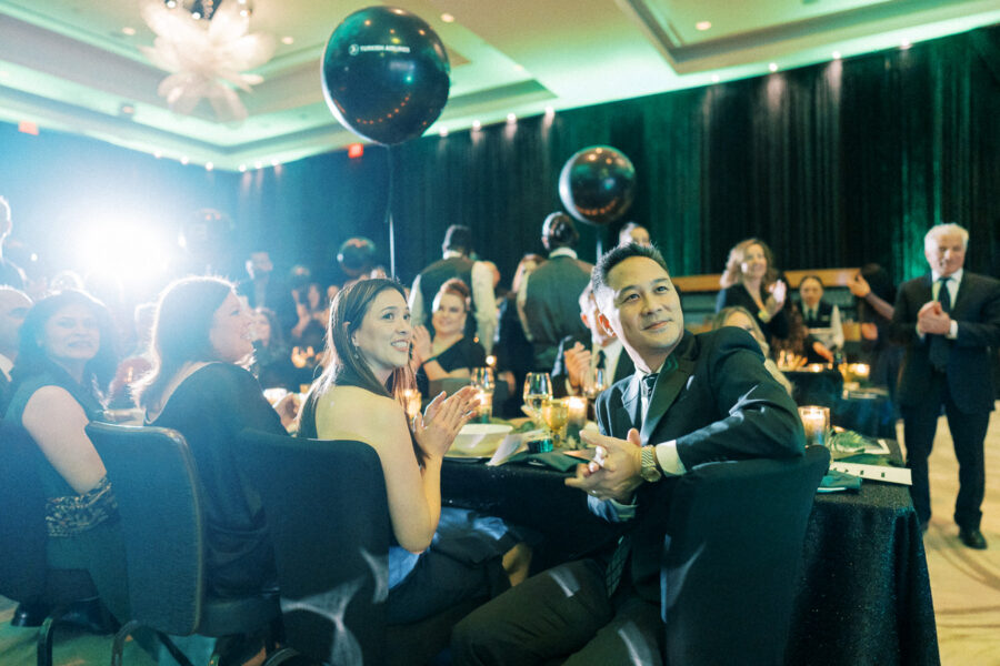 A formal event with elegantly dressed attendees sitting and clapping at round tables adorned with candles and wine glasses, with ambient green lighting and decorative black balloons in the background.