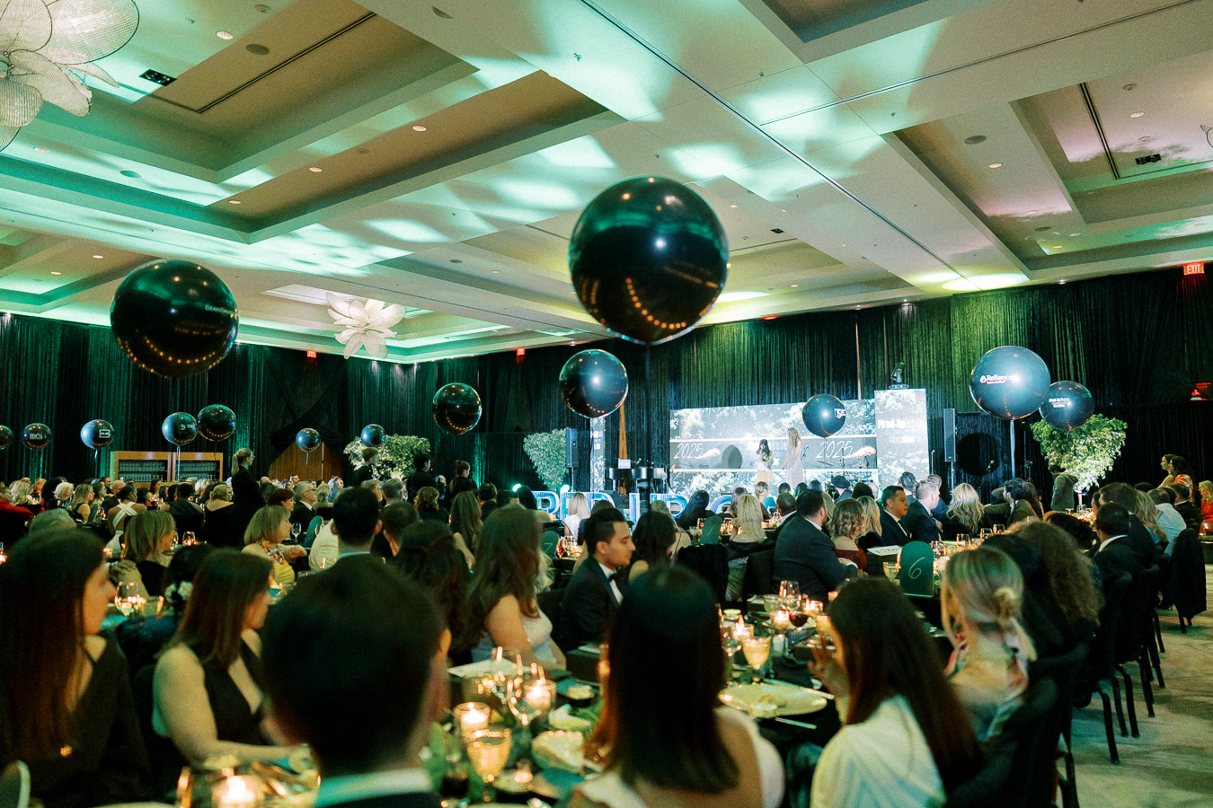 Elegant corporate gala dinner with guests seated at round tables, ambient lighting, and large black balloons decorating a spacious conference hall.