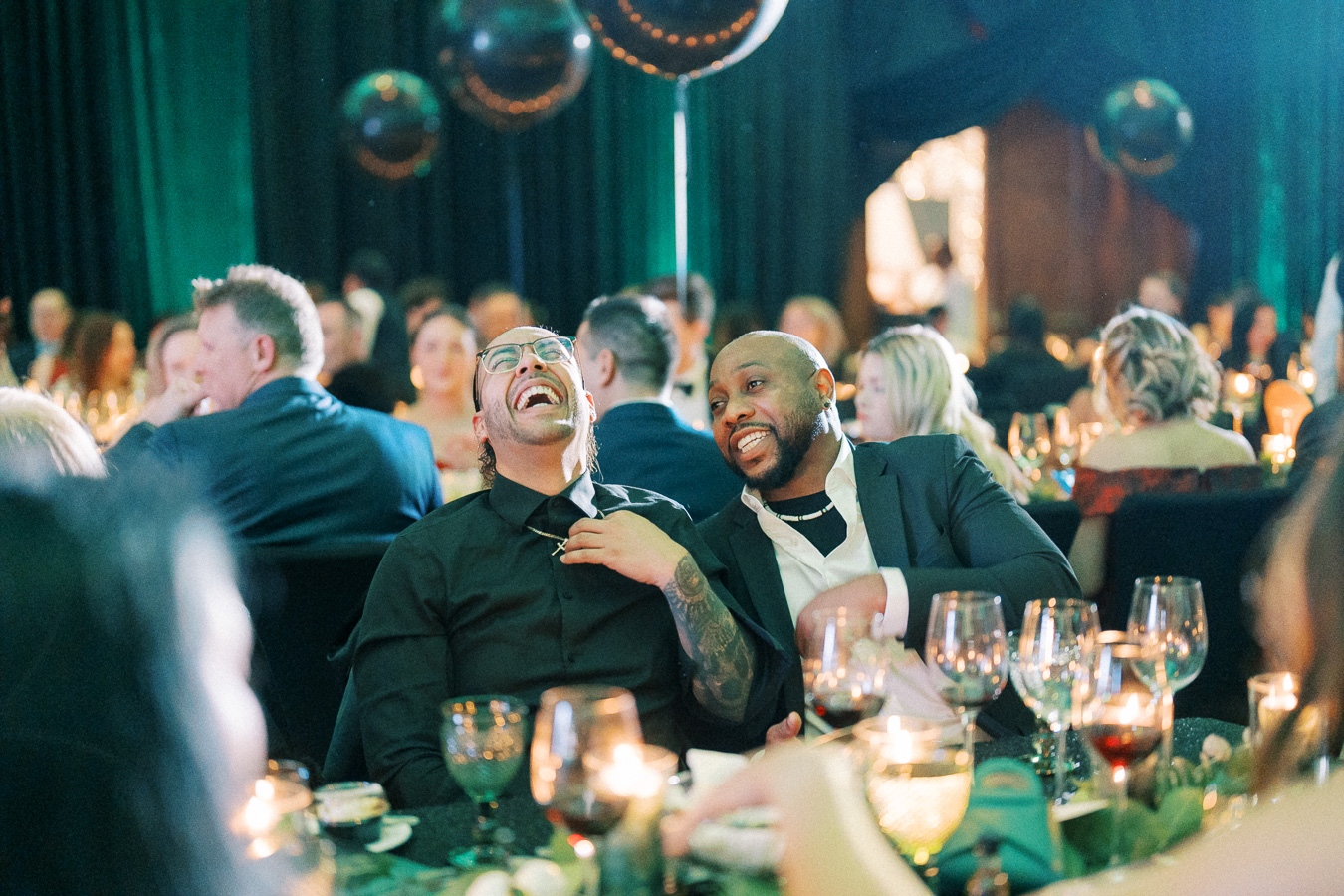 Two men sharing a joyful moment at an elegant event, surrounded by a seated crowd and candlelit tables, creating a lively and celebratory atmosphere.