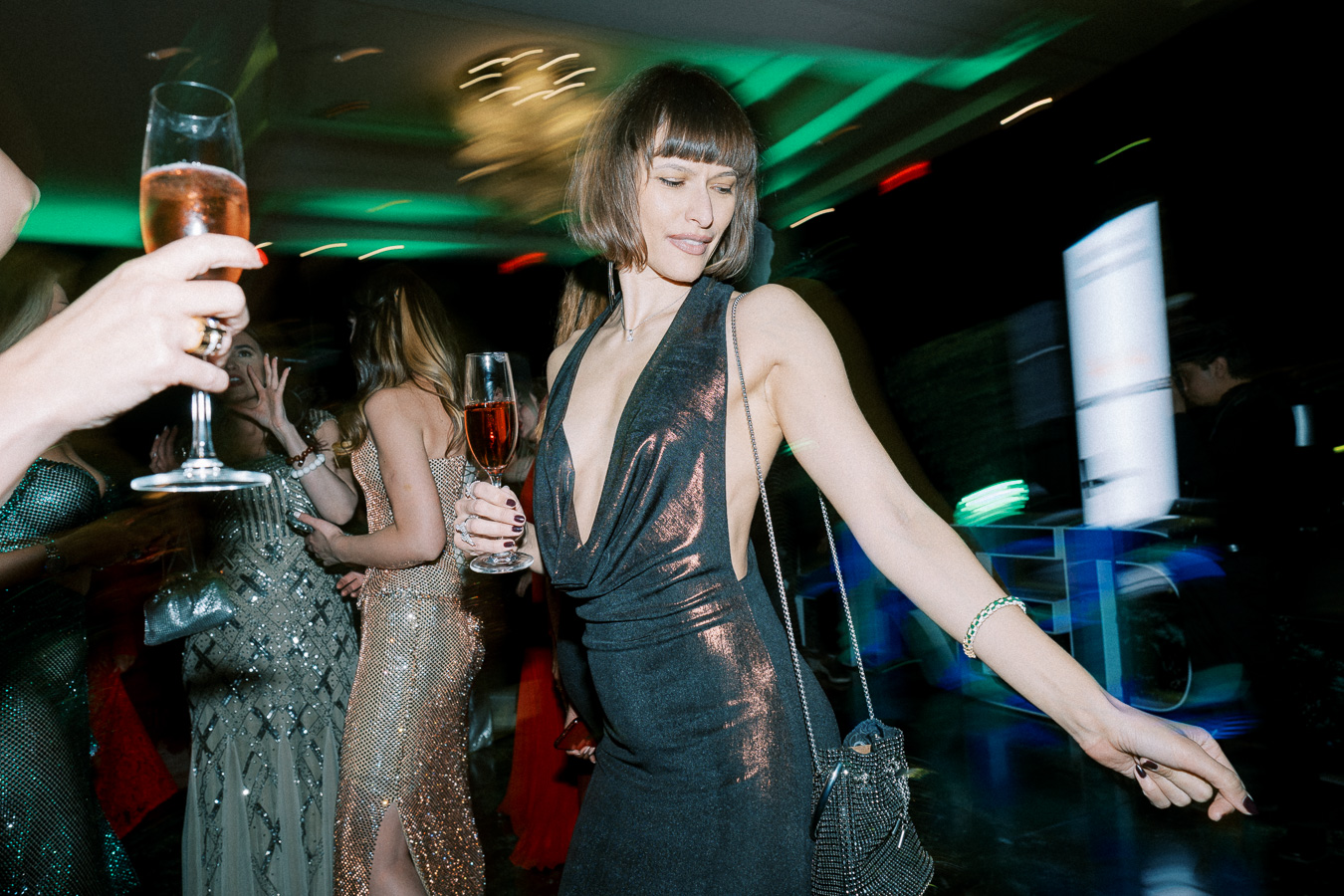 A woman in a stylish dark dress and bob haircut dances at a lively party, holding a glass of champagne. The background is full of colorful lights and elegantly dressed people, creating a vibrant, festive atmosphere.