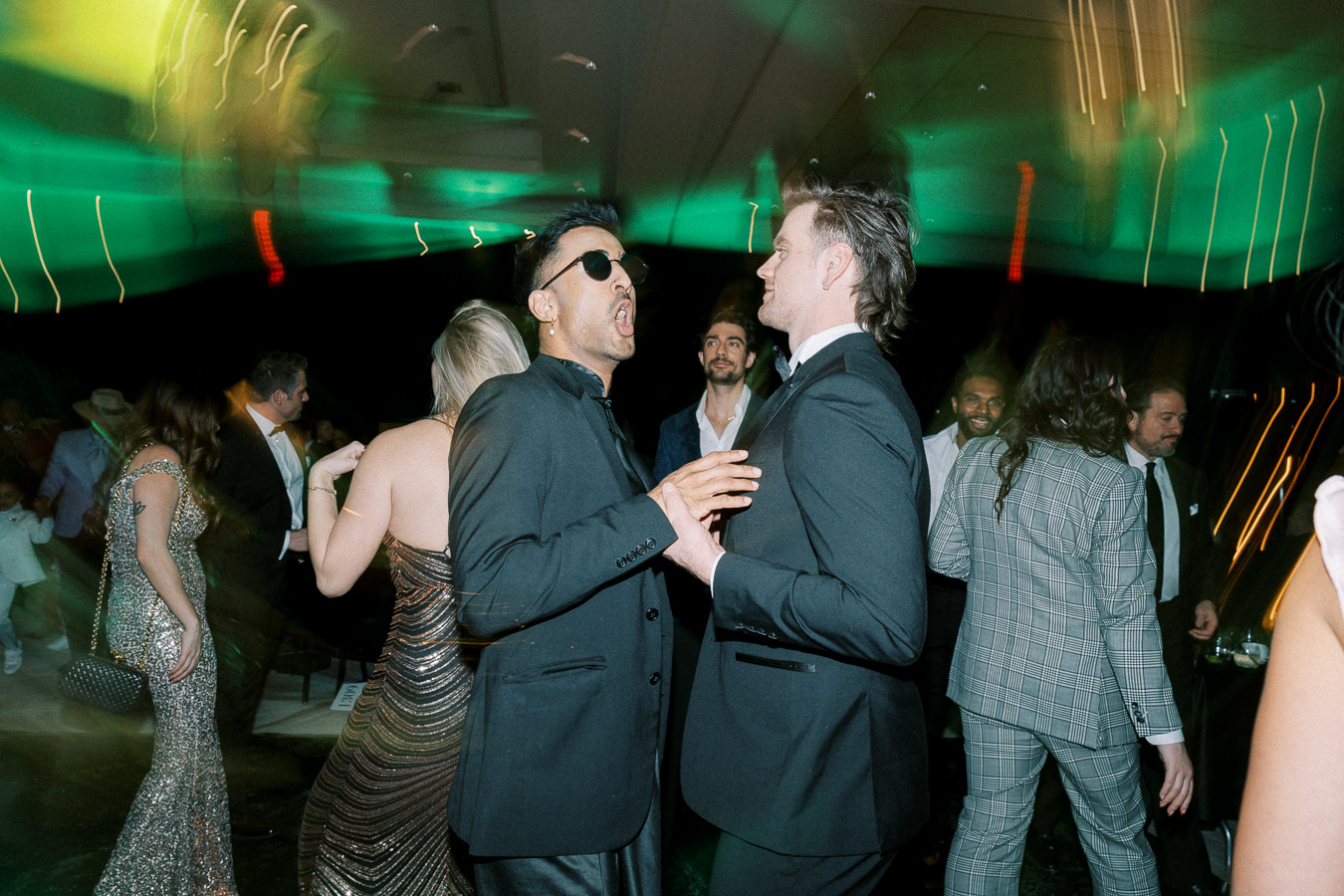 A lively party scene with elegantly dressed people dancing, featuring a well-dressed man in sunglasses animatedly engaging with another well-dressed man. The background is filled with guests enjoying the vibrant atmosphere, enhanced by colorful lighting.
