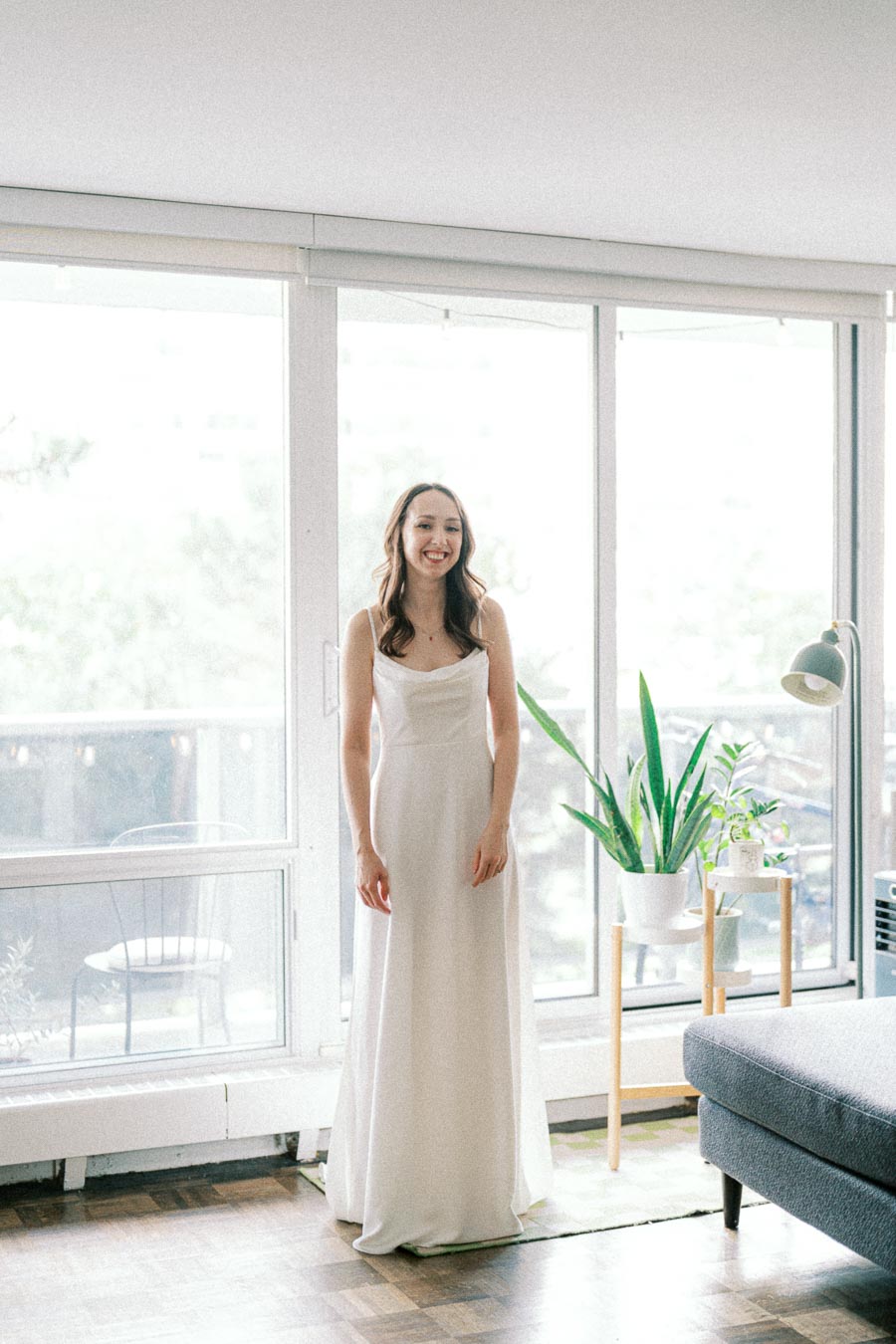 A bride in a white wedding dress standing in a sunlit living room with large windows and a potted plant nearby.