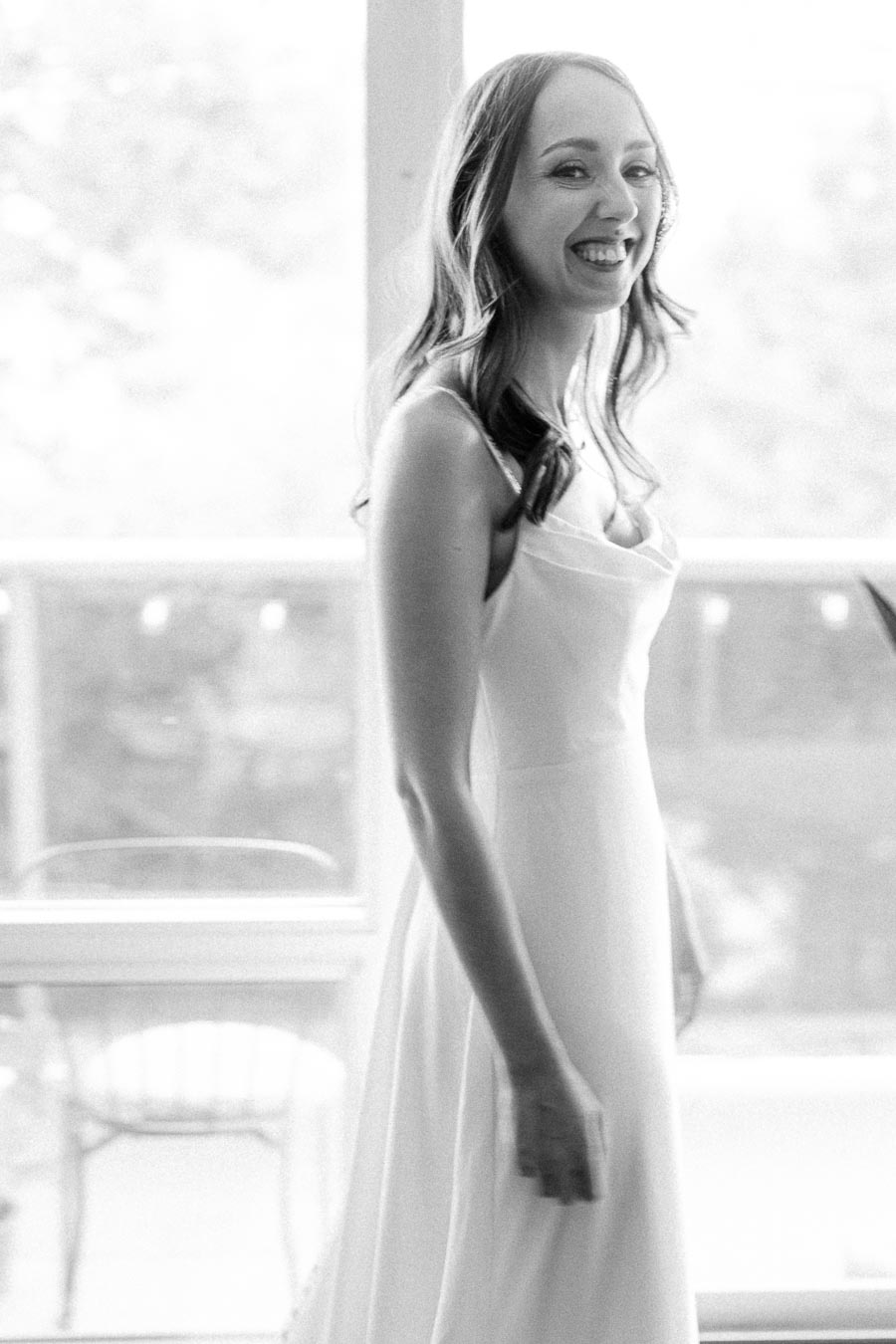 Black and white photo of a smiling woman in a flowing white dress, standing near a bright window, exuding joy and elegance.