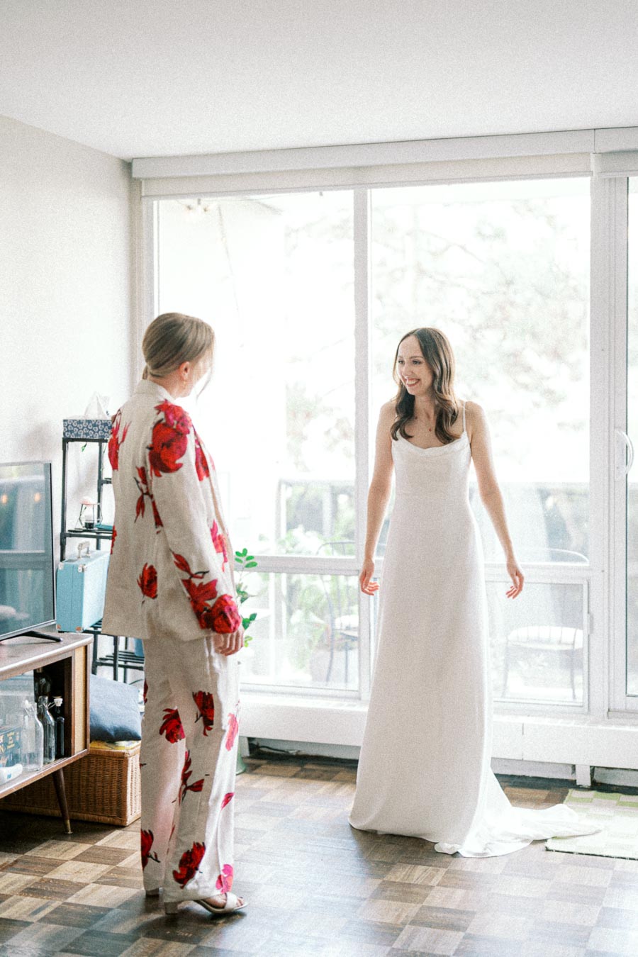 Bride in elegant wedding dress smiling at friend in a floral suit, standing in a bright room with large windows.