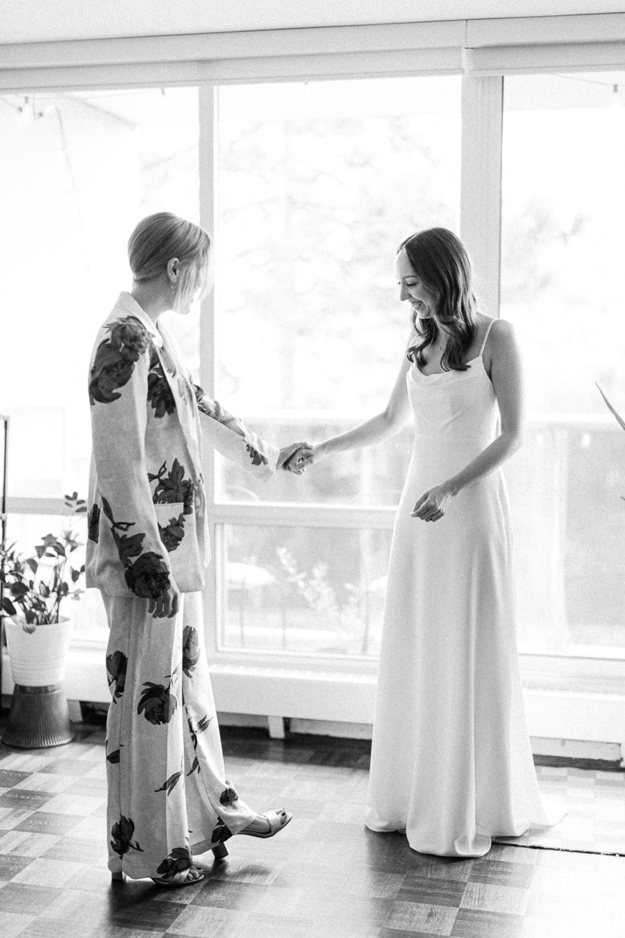 Black and white photo of two individuals holding hands in a bright room, one in a floral suit and the other in a simple white dress, with large windows in the background and plants nearby.