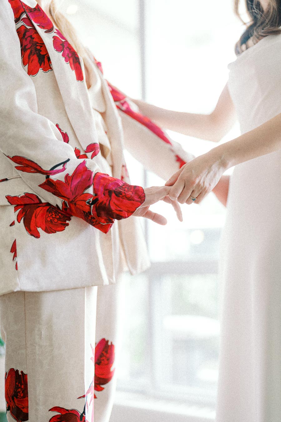 Two people holding hands, one in a white suit with vibrant red floral patterns, and the other in a white dress, with a bright and airy background.