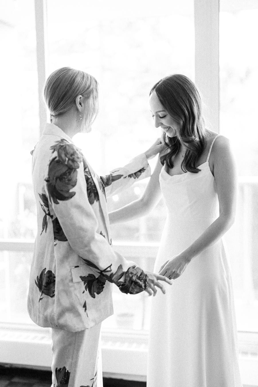 Two women smiling and sharing a joyful moment, one wearing a floral suit and the other in a white dress, in a bright setting.