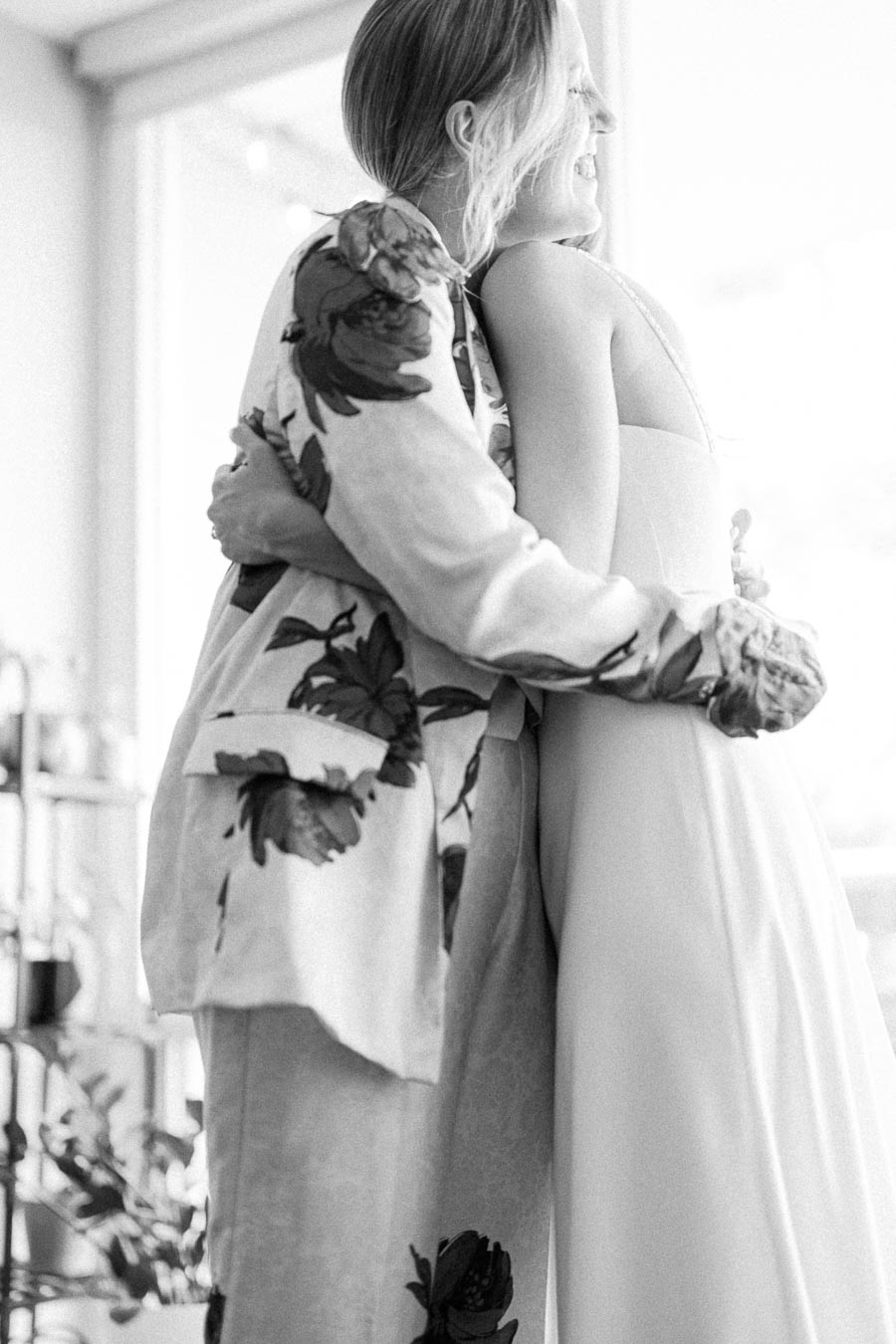 Black and white photo of two people embracing, one wearing a floral blazer and the other in a dress, capturing a joyful and intimate moment indoors.