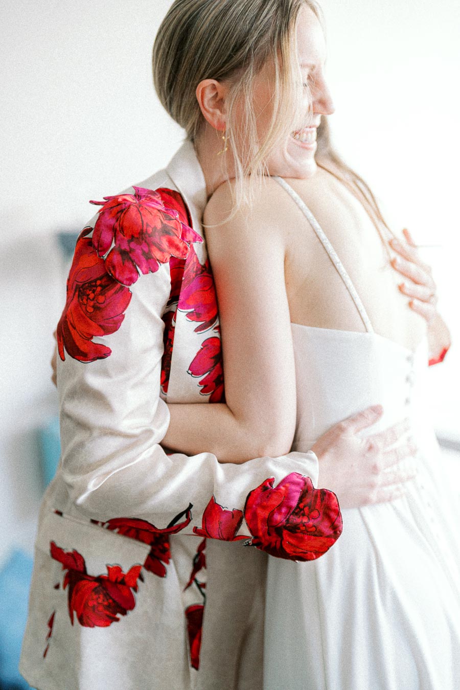 A woman in a floral-patterned jacket embraces another woman in a white dress, both smiling warmly, creating a moment of joy and connection.