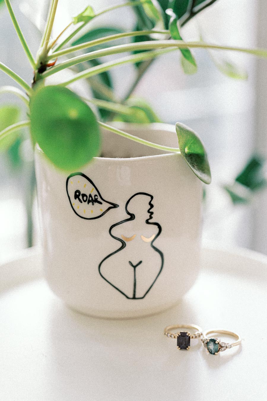 Ceramic mug with abstract art and speech bubble reading roar, next to two elegant gemstone rings on a white surface, accompanied by a leafy green plant.