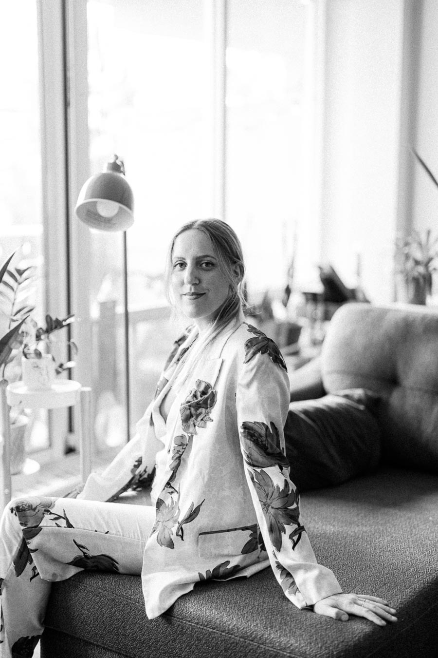 Black and white portrait of a woman sitting on a sofa in a bright room, wearing a stylish floral outfit with plants and a lamp in the background.