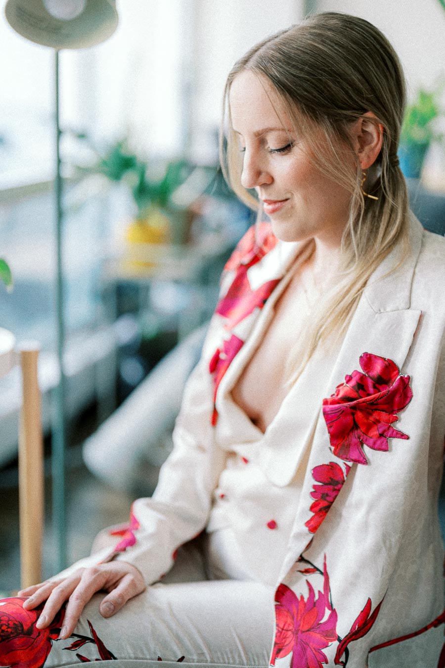 Woman in a stylish white suit adorned with red floral patterns, sitting and looking down thoughtfully.